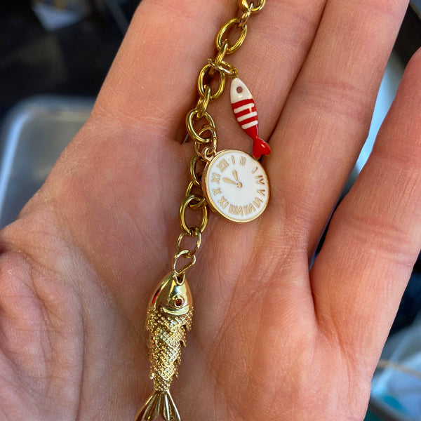 A gold-tone charm bracelet held in a hand, featuring a lobster clasp, a small white clock charm with Roman numerals, a red-and-white striped fishing lure charm, and a detailed gold fish charm.