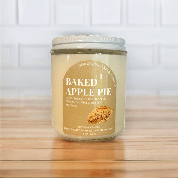 A glass jar candle labeled 'Baked Apple Pie' by Gorgeous Waffle Street, filled with a warm cream-colored wax and resting on a wooden surface against a white brick wall background.