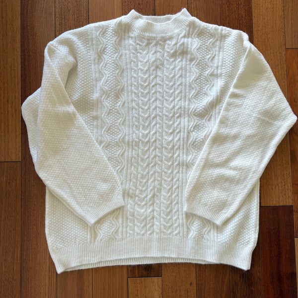 A white cable-knit sweater with a mock neck and ribbed hem, laid flat on a dark brown wooden floor.
