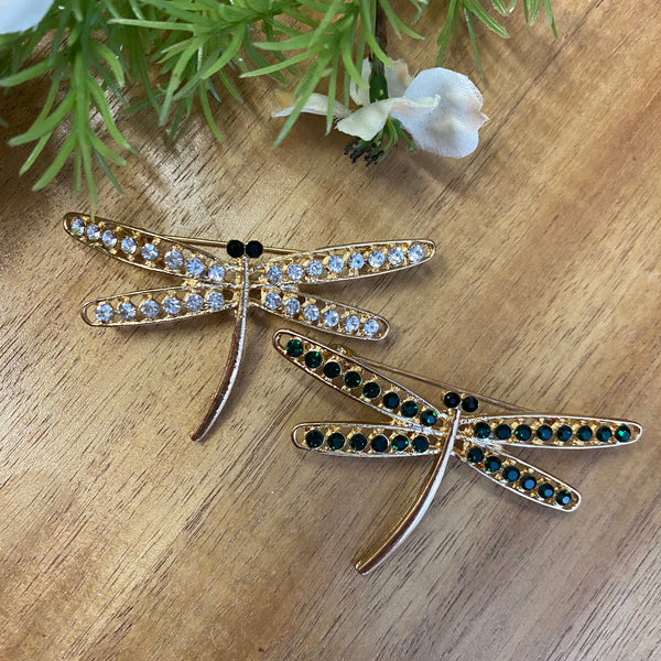 Dragonfly-shaped brooches with gemstones on a wooden surface with greenery.