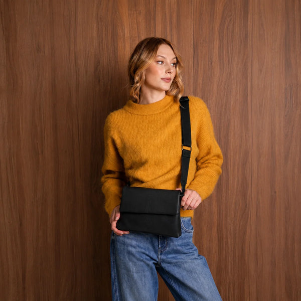 A woman wearing a mustard yellow knit sweater and blue jeans carries a black crossbody messenger bag with a black strap against a dark wood-paneled wall.