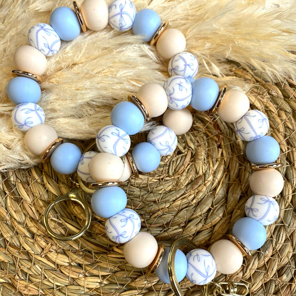 Two beaded bracelets with light blue, cream, and white patterned beads, accented with gold spacer rings and gold-tone lobster clasps, displayed on a woven straw mat with a beige faux fur background.