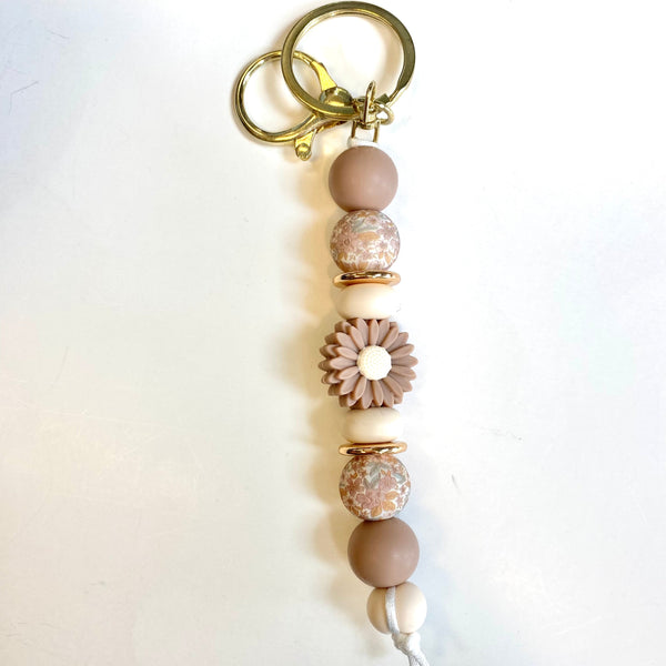 A handmade beaded keychain with a gold-tone split ring and clasp, featuring matte brown and cream beads, patterned beads, a flower-shaped bead, and a white cord tassel on a white background.