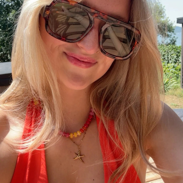 A close-up selfie of a blonde woman wearing tortoiseshell sunglasses and a bright orange-red bikini top, outdoors on a wooden deck with green foliage in the background.