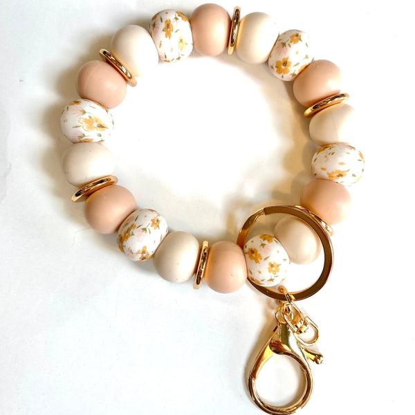 A beaded bracelet with pastel peach, cream, and white beads with gold foil details, separated by gold spacer rings, and finished with a gold lobster clasp and key ring charm, shown on a white background.