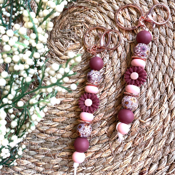 Two beaded keychains with copper-toned split rings, featuring matte burgundy and pink beads, floral bead accents, and textured purple marbled beads, displayed on a woven straw mat with baby's breath flowers.