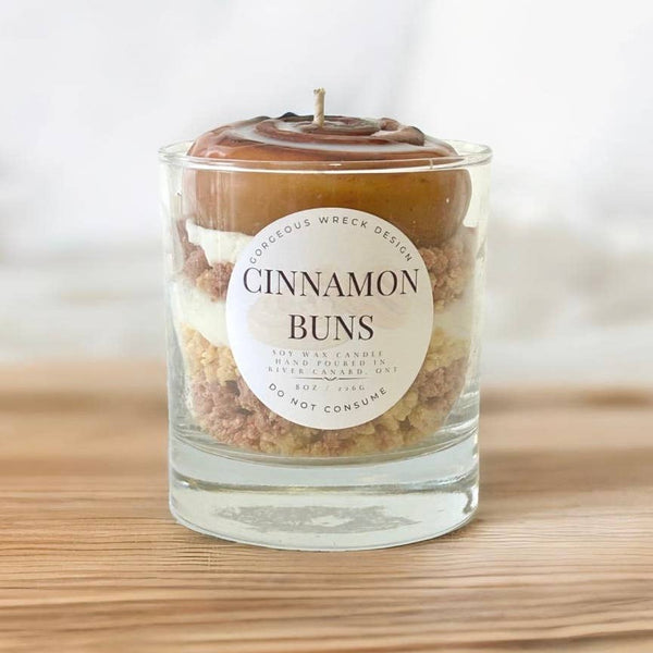 Candle in a glass jar labeled 'Cinnamon Buns' on a wooden surface with a white background