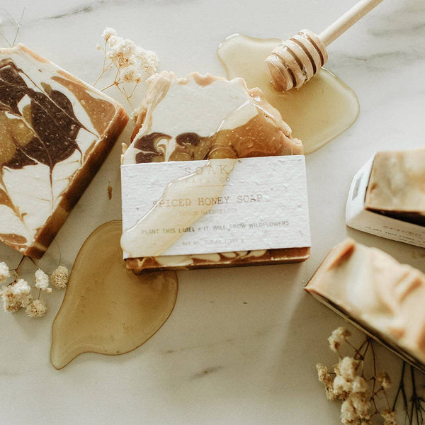 A styled flat-lay of artisanal soap bars on a light marble surface, with honey drizzles, a wooden honey dipper, and small dried flowers. The central product is a bar of soap wrapped in a white paper band labeled 'SOAK Spiced Honey Soap'.