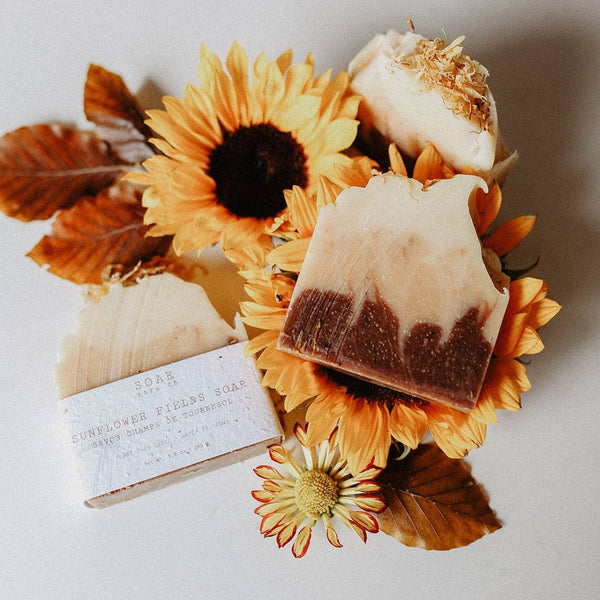Three handmade artisan soaps arranged with dried sunflowers and autumn leaves on a light gray background. One soap has a white paper label reading 'SUNFLOWER RUMORS SOAP'.