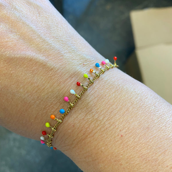A close-up of a person's wrist wearing a delicate gold chain bracelet adorned with small colorful bead drops in red, orange, yellow, green, blue, pink, and white.