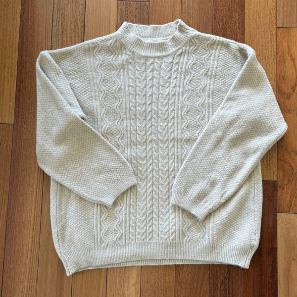 A cream-colored cable-knit sweater with a mock neck, laid flat on a wooden plank floor.