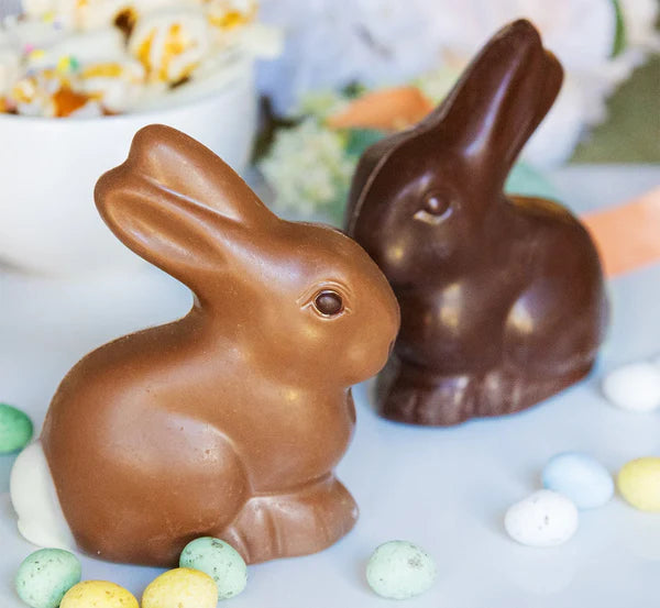 Two chocolate bunnies on a table with Easter eggs and candy.