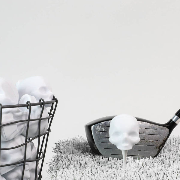 Golf club with a golf ball on grass next to a wire basket with golf balls against a white background