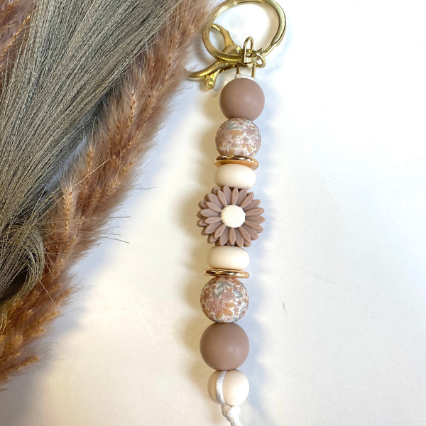 A boho-style beaded keychain charm with a brass key ring, featuring matte and textured brown beads, a brown flower bead with a cream center, and a small white cord tie, styled next to dried pampas grass on a white background.