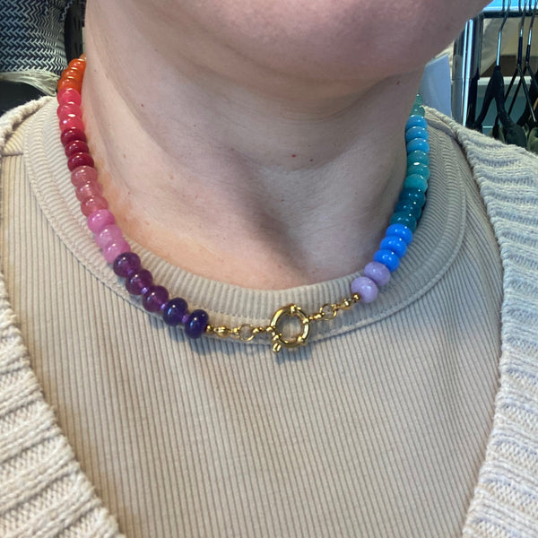 A close-up of a person wearing a beaded choker necklace with a gold clasp. The necklace features a gradient of beads transitioning through red, orange, pink, purple, blue, and green, worn over a ribbed beige top.