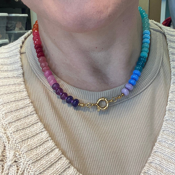 A close-up of a person wearing a double-strand beaded necklace with a rainbow gradient from red and pink to purple, blue, and green, accented by a gold chain with a circular interlocking ring clasp.