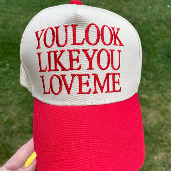 A red and off-white trucker-style baseball cap held by a hand against a green grass background. The front panel is off-white with red embroidered text reading 'YOU LOOK LIKE YOU LOVEME'.