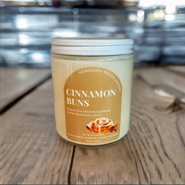 A jar candle labeled 'Cinnamon Buns' sits on a wooden tabletop with a blurred bakery-themed background. The candle has creamy off-white wax in a clear glass jar with a white lid and a tan label featuring a cinnamon roll illustration.