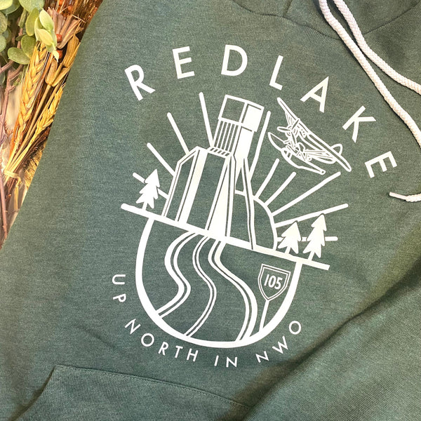 A close-up of a dark heather green hoodie featuring a white graphic print on the chest. The graphic includes the text 'REDLAKE' and 'UP NORTH IN NWO' surrounding an illustration of a mountain, roads, trees, and a plane.