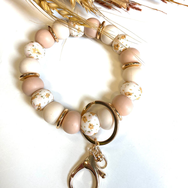 A beaded keychain bracelet featuring pastel pink, cream, and white beads with gold metallic accents, including a large gold ring and lobster clasp, styled with dried wheat on a white background.