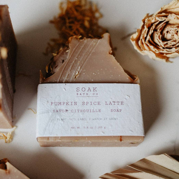 A bar of Soap Bath Co. Pumpkin Spice Latte savour citrouille soap, wrapped in a textured white paper label, displayed on a light beige surface with dried botanical elements.