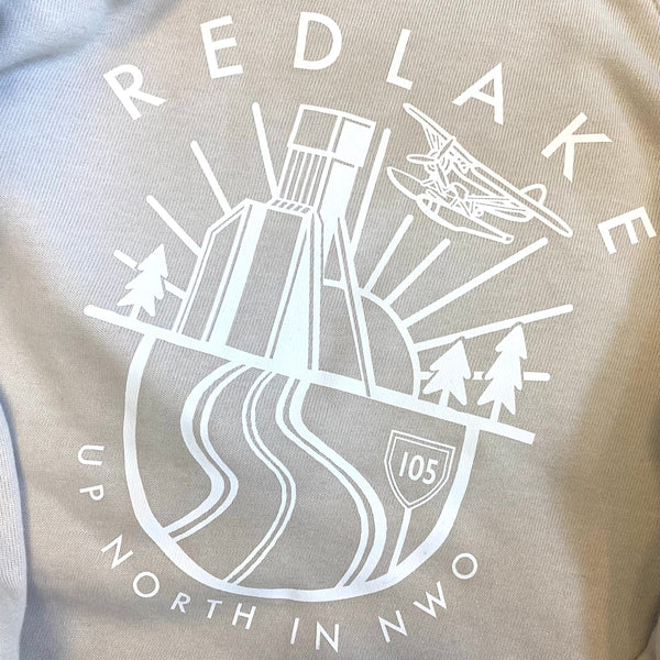 Close-up of a beige or light taupe sweatshirt with a large white graphic print on the front. The graphic features the word 'REDLAKE' arched over an illustration of a building, trees, a car, and winding roads, with 'UP NORTH IN NWO' curved below.