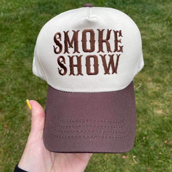 A two-tone baseball cap held by a hand outdoors on a grassy lawn. The cap features an off-white crown with dark brown embroidered text reading 'SMOKE SHOW' and a dark brown visor.