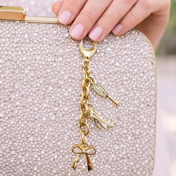 A close-up of a hand holding a sparkling silver clutch bag with a gold-tone charm keychain featuring a bow, key, and cigarette lighter details.