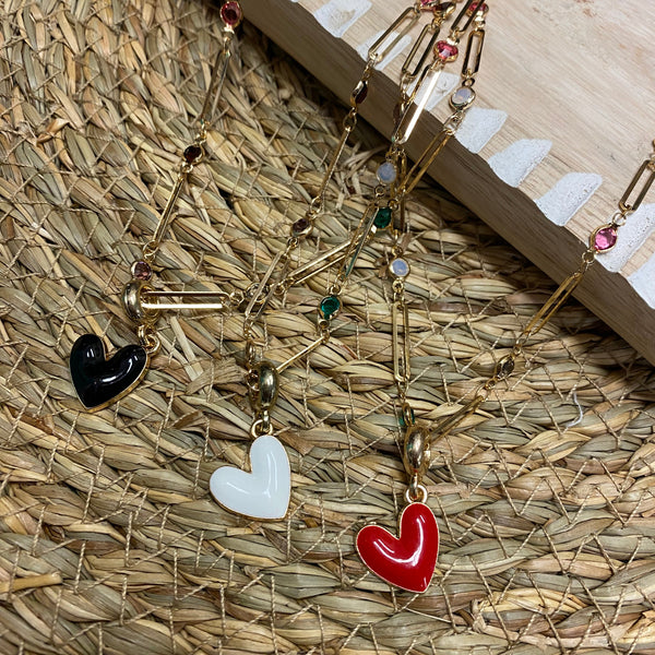 A gold-tone beaded necklace with heart-shaped charms in red, white, and black, displayed on a woven straw mat.