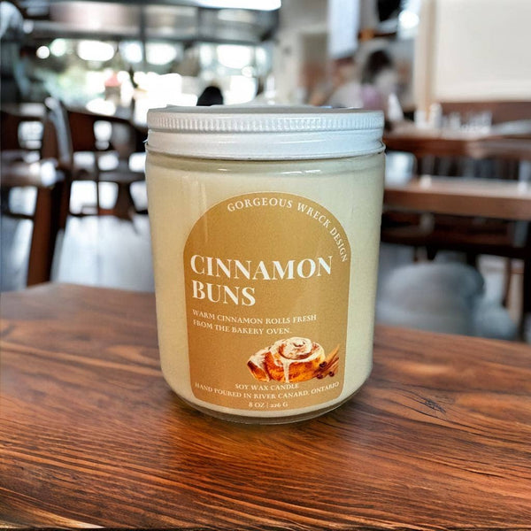A jar candle labeled 'Cinnamon Buns' by Gorgeous Wreath Designs sits on a wooden table in a softly blurred cafe setting.