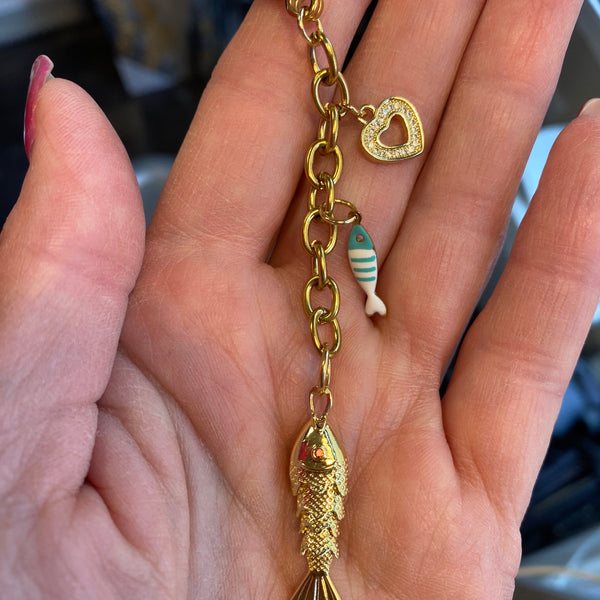 A gold-tone chain bracelet with a fish charm and a small striped bead, held in a person's hand.