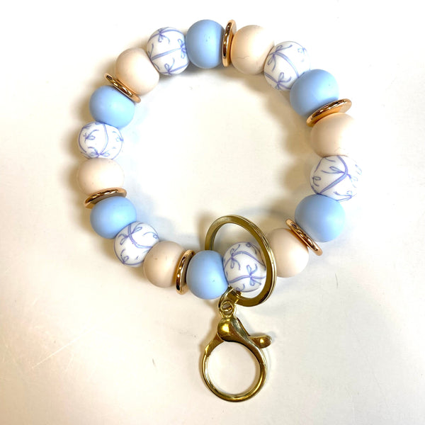 A beaded bracelet with light blue, cream, and white patterned beads, separated by small gold spacer rings, with a gold lobster clasp charm, shown on a plain white background.