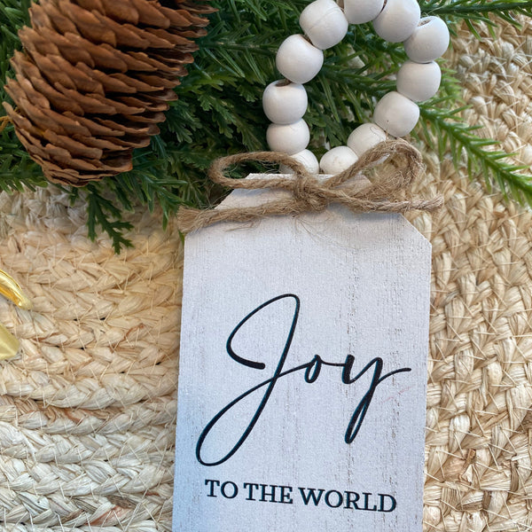 Decorative tag with 'Joy to the World' text on a textured surface with greenery and beads.