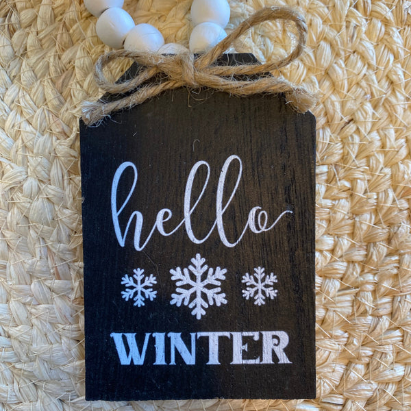 Small wooden sign with 'hello winter' text and snowflake designs on a textured surface.