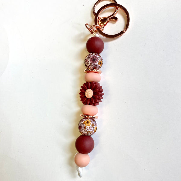 A beaded keychain with rose gold metal key rings, featuring matte burgundy and peach beads, a dark burgundy flower bead, and two patterned glass beads on a plain white background.