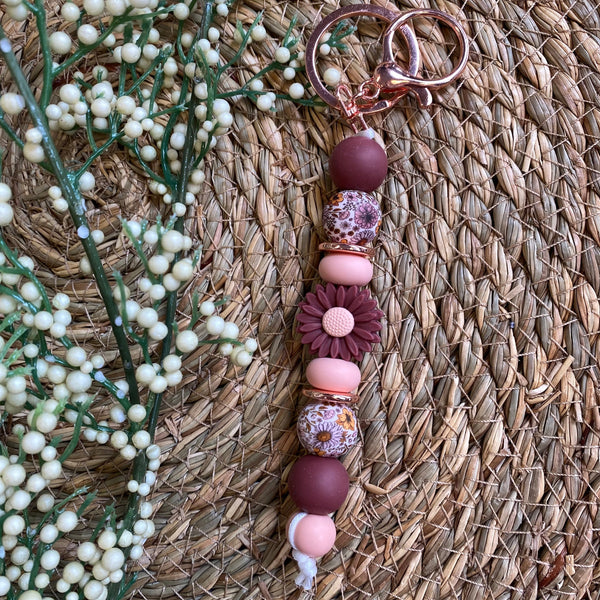 A beaded keychain with rose gold metal rings, featuring matte burgundy and pink beads, patterned glass beads, and a small flower bead, styled on a woven straw mat with baby's breath flowers.