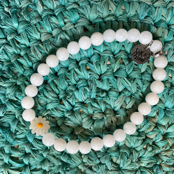 A beaded bracelet made of white round beads, featuring a small white daisy charm and a silver-toned round charm engraved with 'LOVE', displayed on a textured teal woven mat.