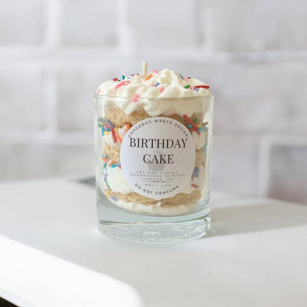 Birthday cake-themed candle in a glass jar with a label on a white surface.