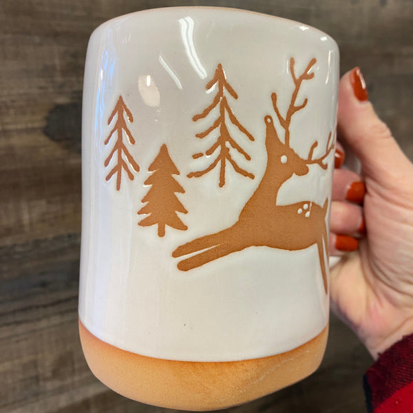 White mug with brown reindeer and tree design held by a hand on a wooden surface