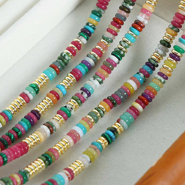 A close-up of multiple strands of colorful beaded jewelry laid diagonally across a light neutral surface. The beads include pink, turquoise, green, white, yellow, purple, and metallic gold spacer rings.