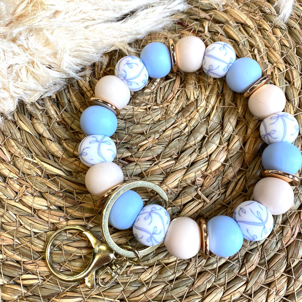 A beaded bracelet with light blue, cream, and white patterned beads, accented with gold-toned spacers and a gold clasp, displayed on a woven straw mat with a cream faux fur accent.