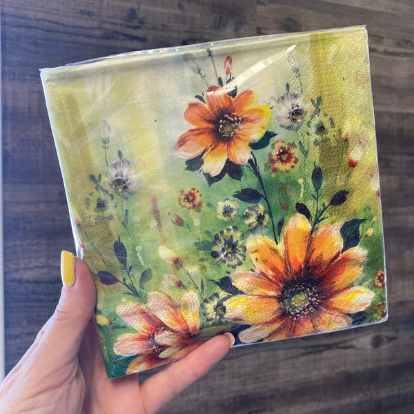 A hand with yellow nail polish holds a square paper napkin featuring a watercolor-style floral design with orange and yellow flowers on a green background, set against a dark wood surface.