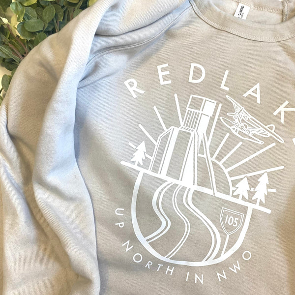 A light cream/ivory crewneck sweatshirt with a white graphic print on the front reading 'RED LAKE UP NORTH IN NWO' around an illustration of a mine shaft, trees, sunset, plane and road.