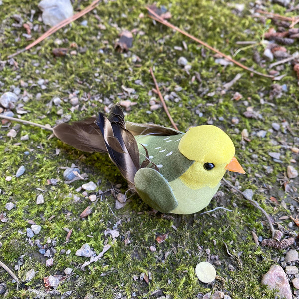 A small artificial bird figurine with a bright yellow head, olive green back, and brown wings, placed on a mossy ground with small stones and pine needles.