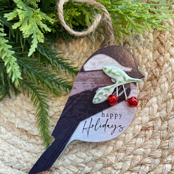 Decorative bird ornament with 'happy holidays' text on a woven surface with greenery.
