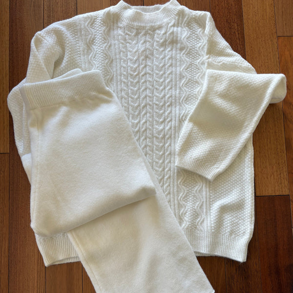 A folded white knit sweater with textured cable and wavy knit patterns, laid flat on a brown wooden floor with plain knit light cream pants and elastic waist sit on top.
