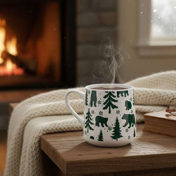 A white ceramic mug with a green winter forest pattern of trees, bears, and snowflakes sits on a wooden table in a cozy living room setting with a burning fireplace in the background.