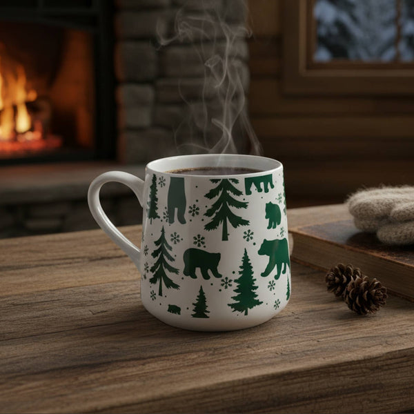 A white ceramic mug with a green forest and bear pattern sits on a rustic wooden table in a cozy cabin setting, with a burning fireplace in the background and a pair of cream knit gloves resting on a book nearby.