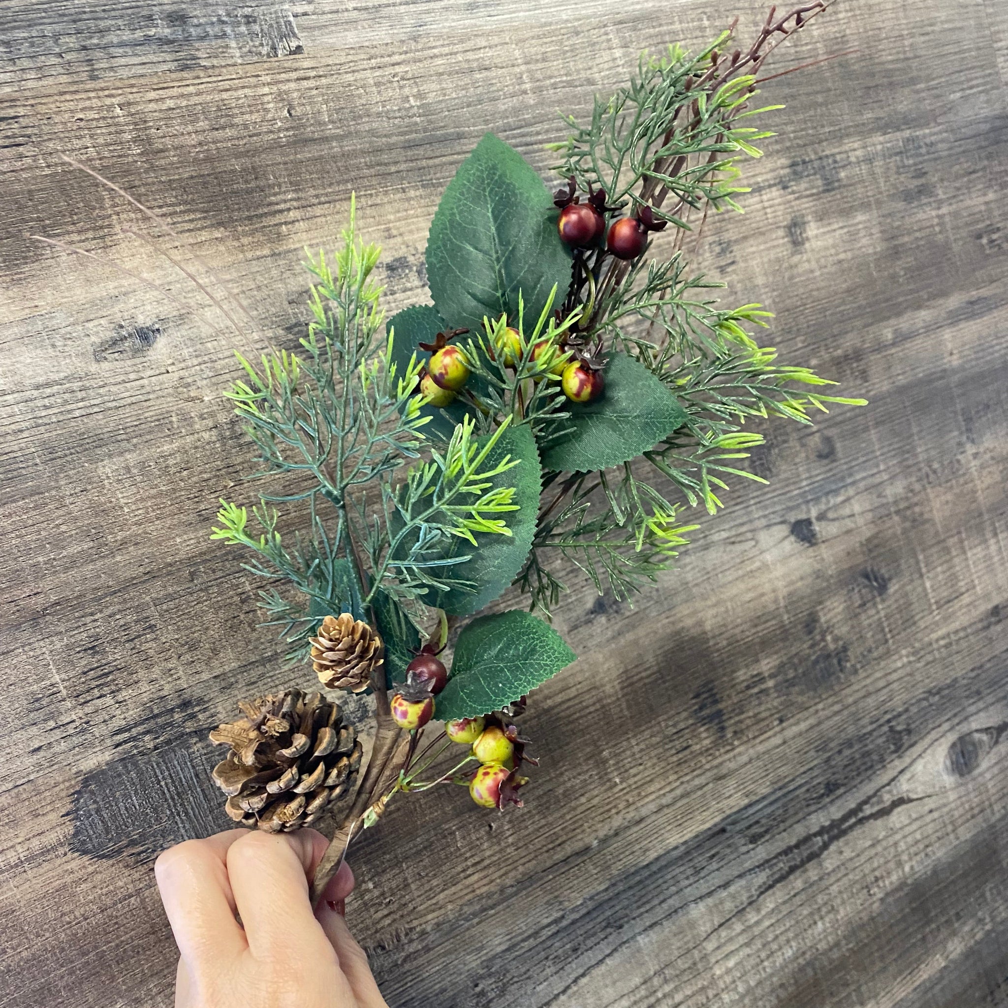 Hand arranging a small bouquet of greenery and pinecones on a wooden surface