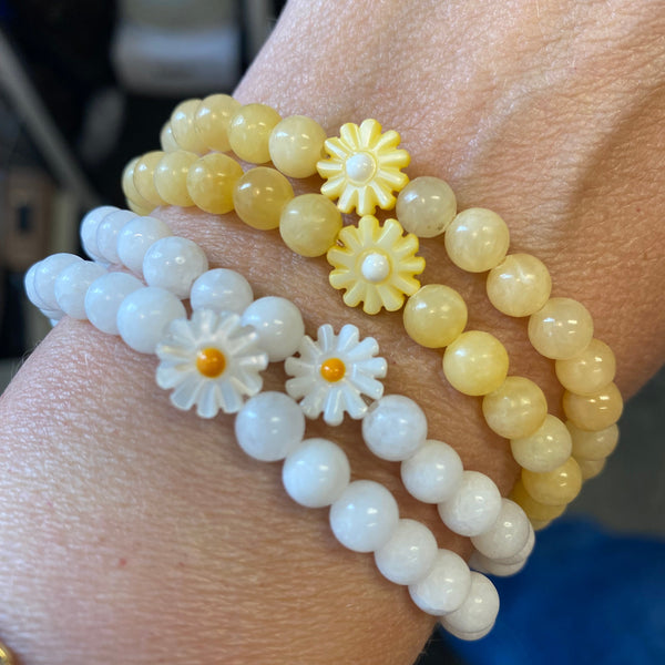 Two beaded bracelets worn on a wrist: one in pale yellow and one in white, both featuring daisy flower charms.